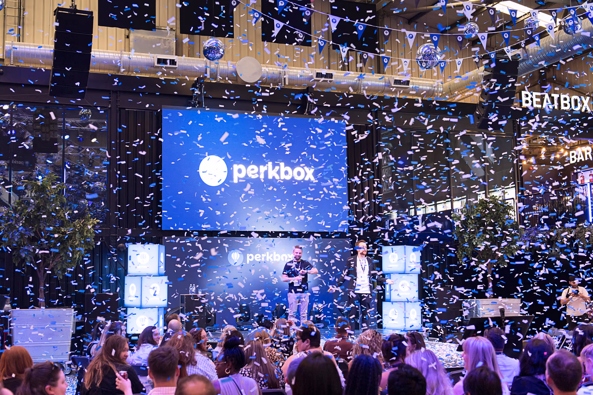 Confetti bursting over a crowd in front of a large branded screen at a company day