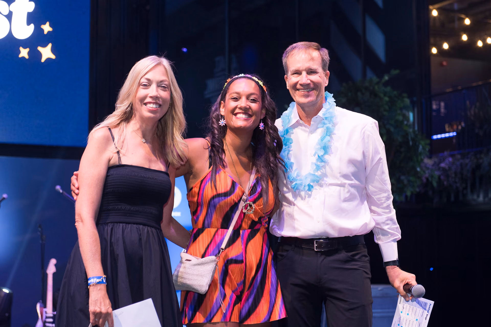 Three colleagues posing together on stage, one wearing a flower garland, at a company celebration