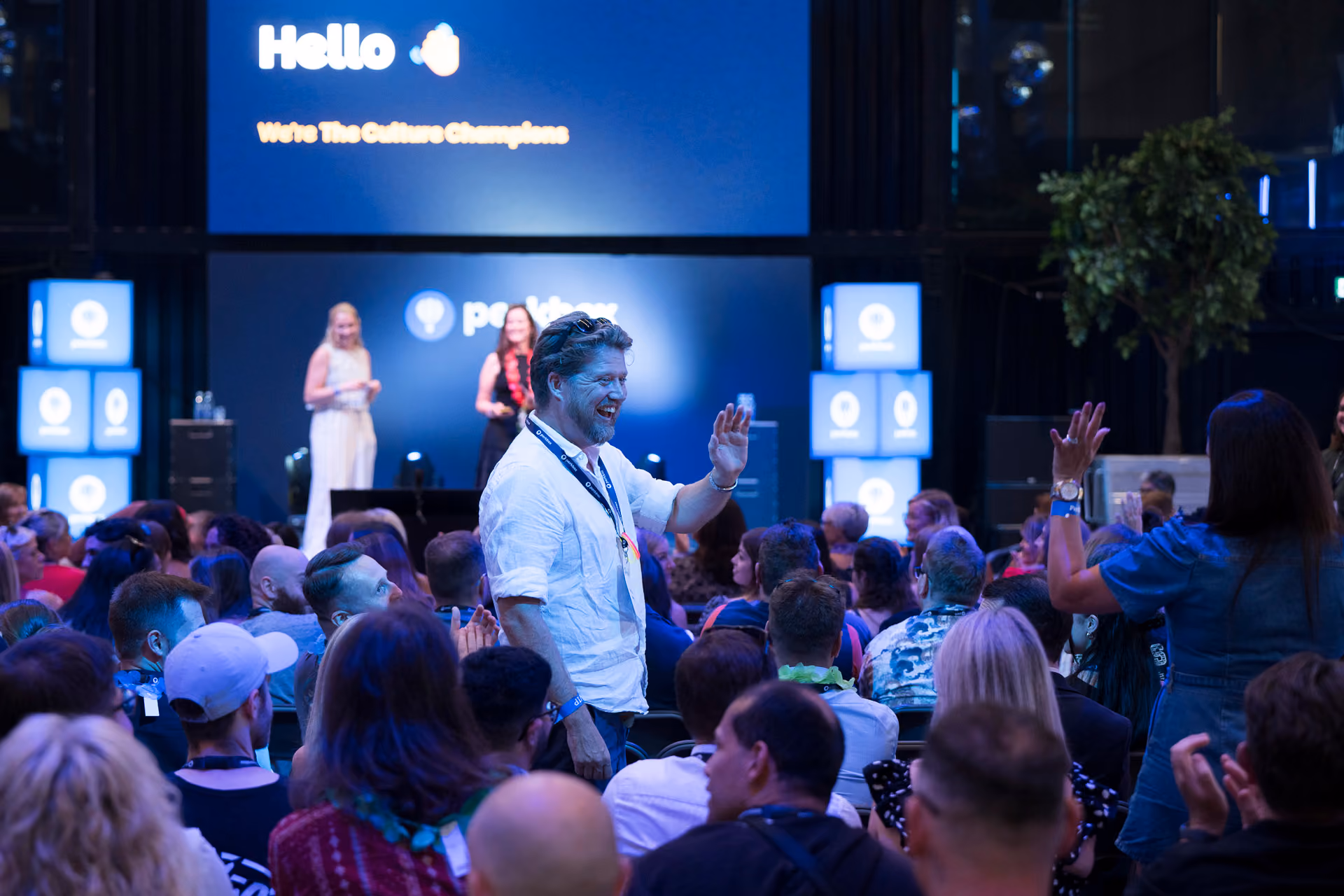 An attendee standing and waving among a seated audience while presenters speak on stage