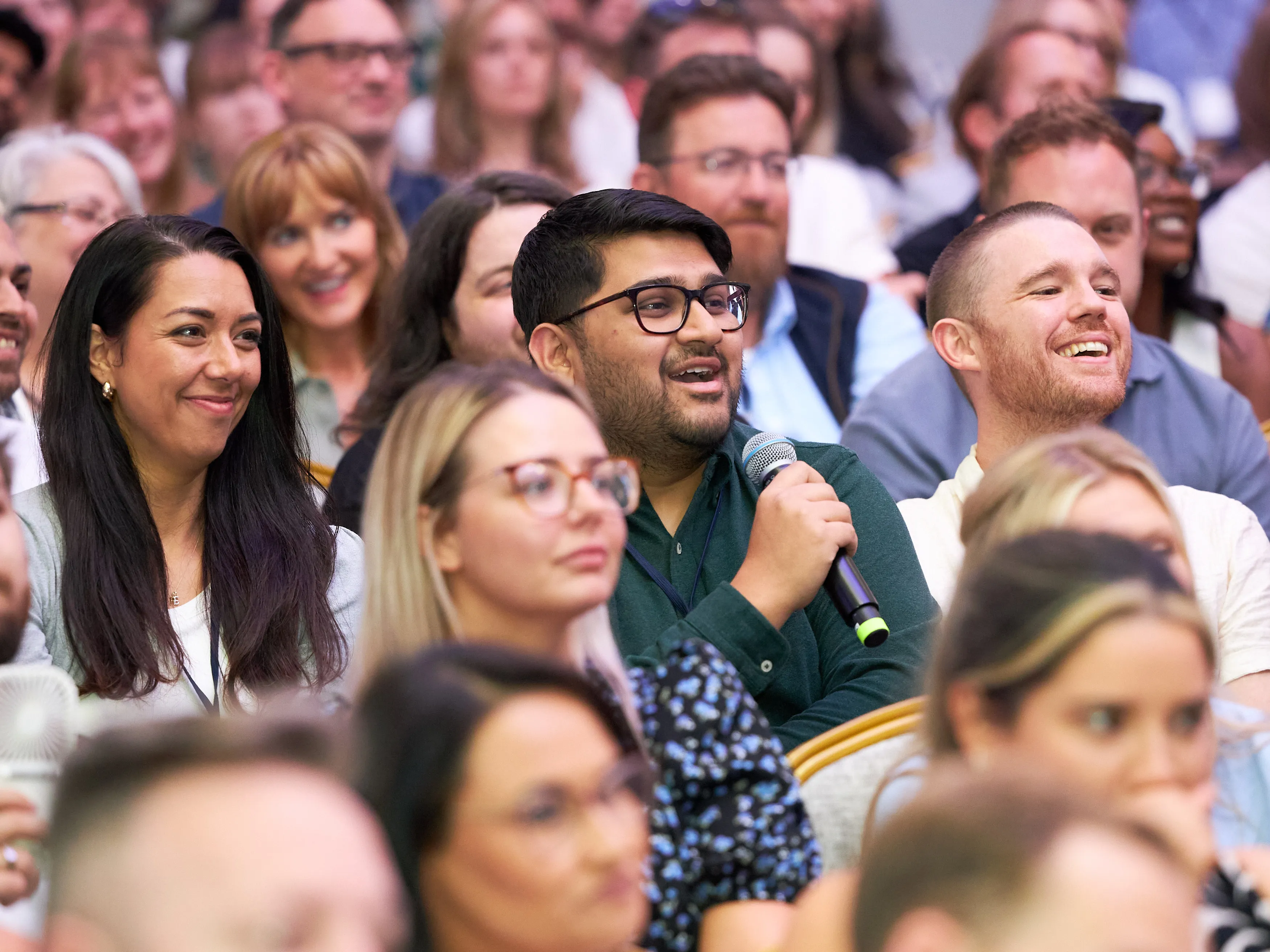 An attendee speaking into a microphone among a smiling, packed conference audience