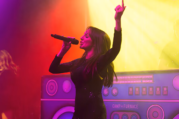 A singer performing under coloured lights beside a giant boombox set at a company party