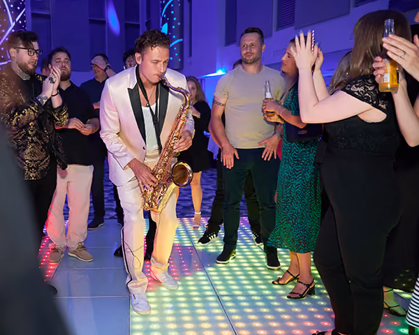 A saxophonist playing on a lit dance floor surrounded by guests at a company party