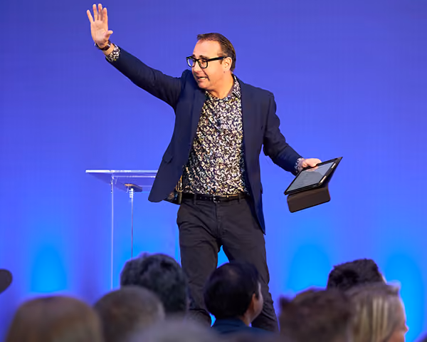 A host waving to the audience from the stage beside a lectern at a conference