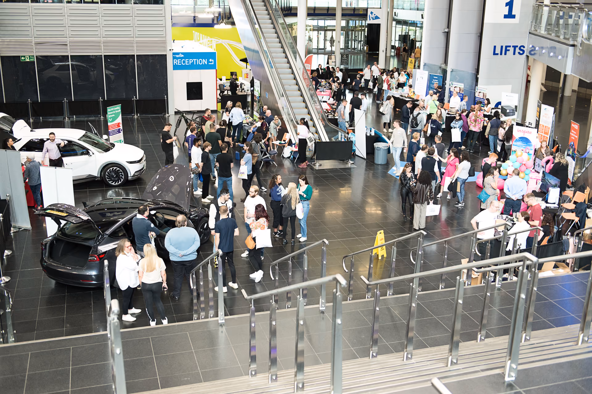 Attendees filling an exhibition hall foyer with cars on display at a corporate event