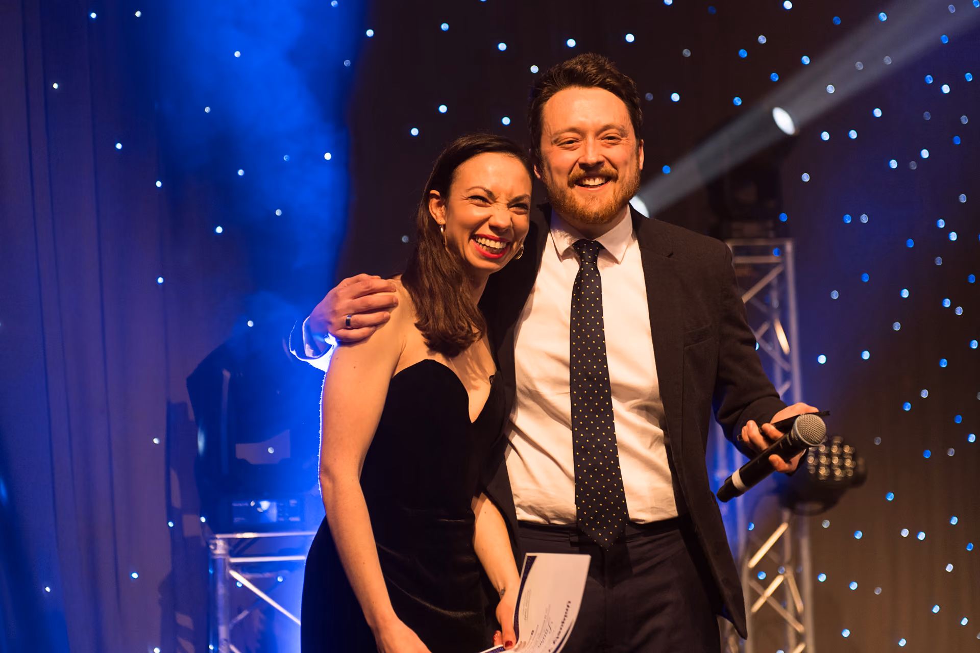 Two hosts with a microphone on stage against a starlit backdrop at an awards evening