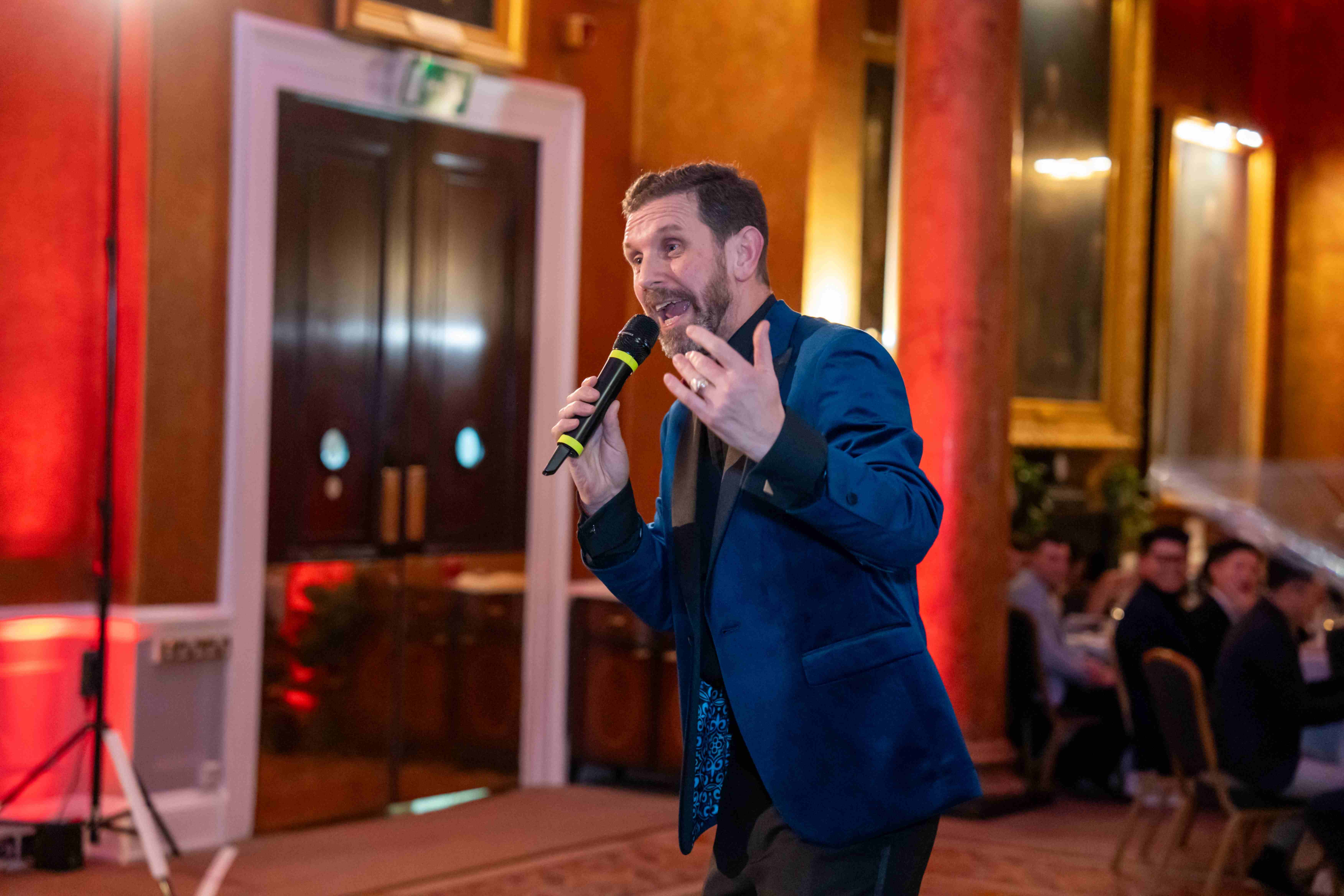 A compere in a blue velvet jacket speaking to seated guests at an awards night
