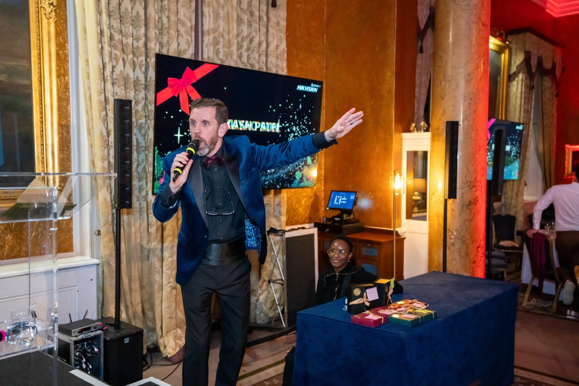 A host with arms outstretched presenting beside a screen at a Christmas awards evening