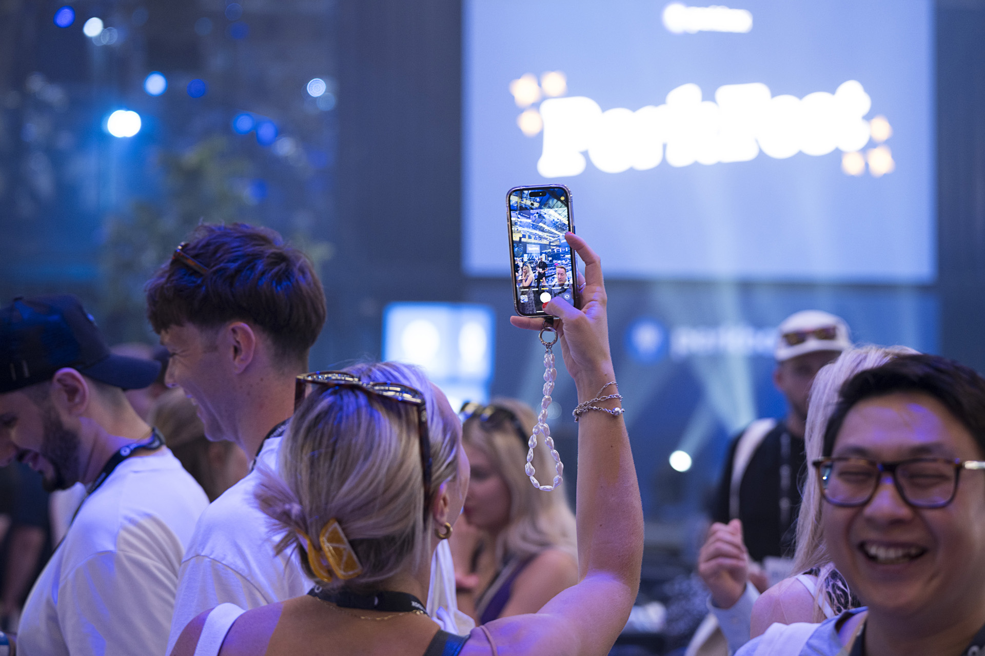An attendee raising a phone to photograph the stage while colleagues laugh nearby