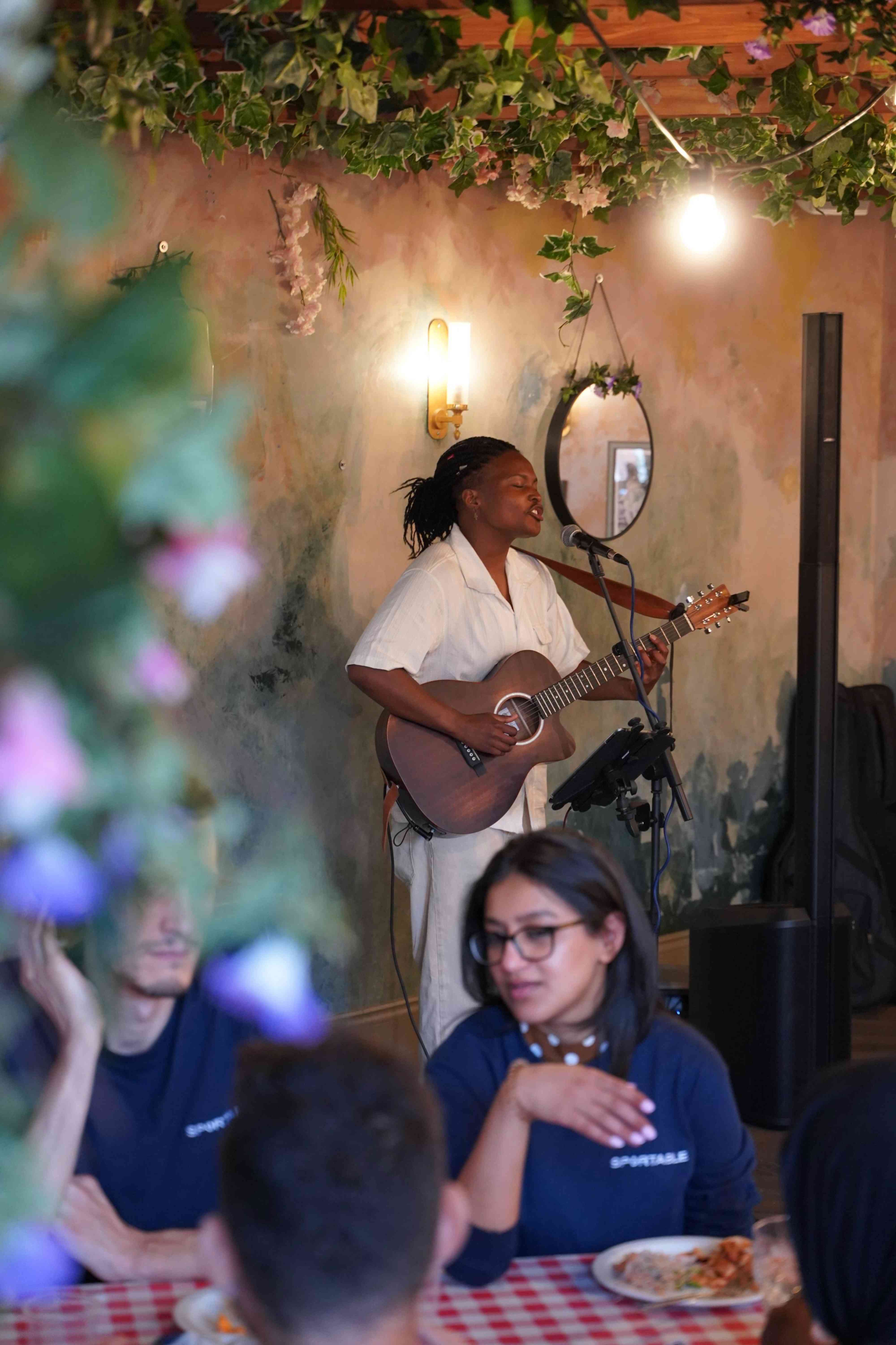 A musician playing acoustic guitar beside diners in a plant-filled restaurant corner