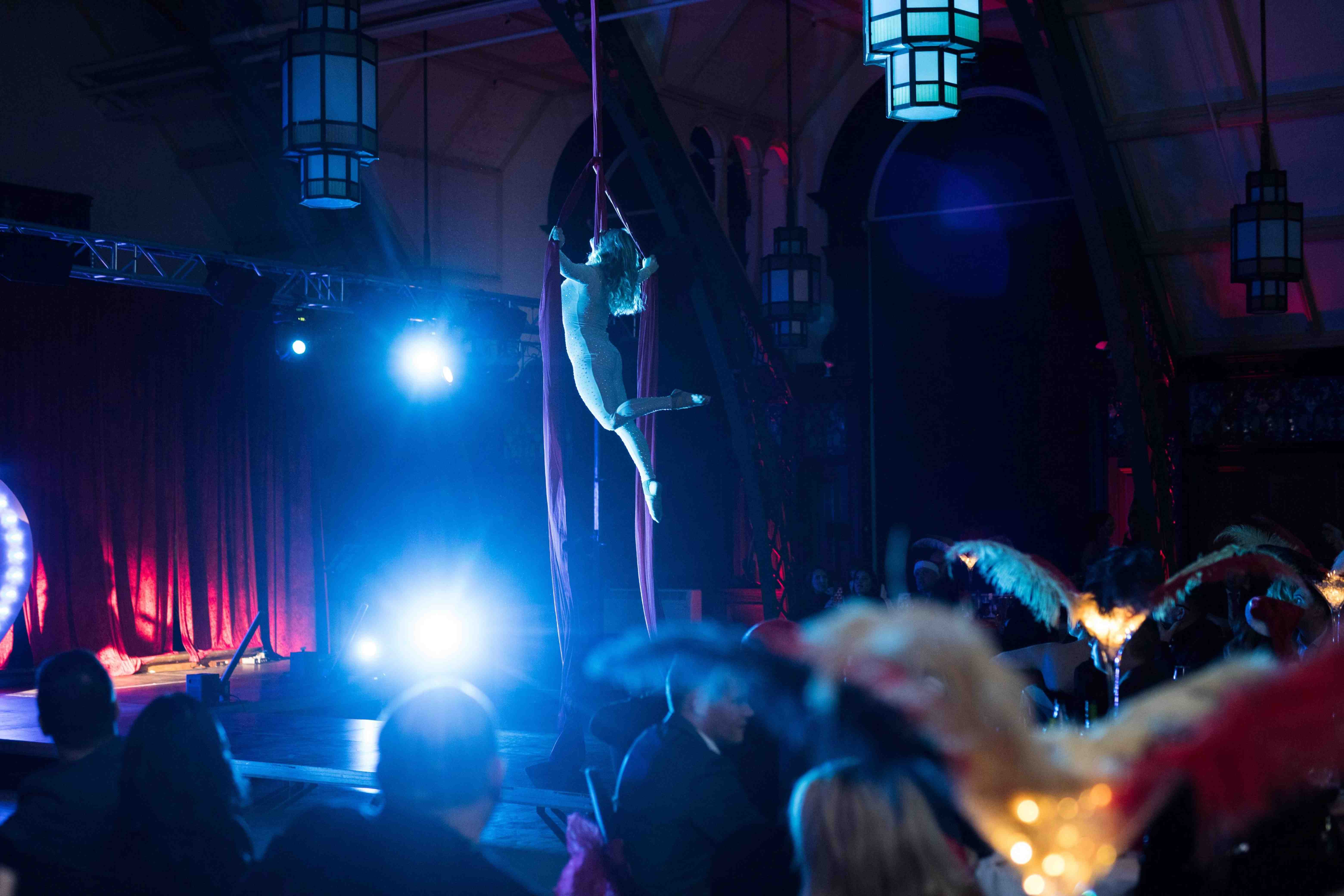Aerial artists suspended above a blue-lit hall as guests in feathered costumes watch