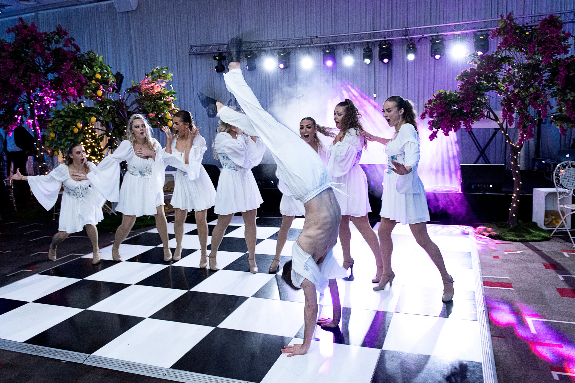 A dance troupe in white costumes performing on a chequered floor beneath floral arches