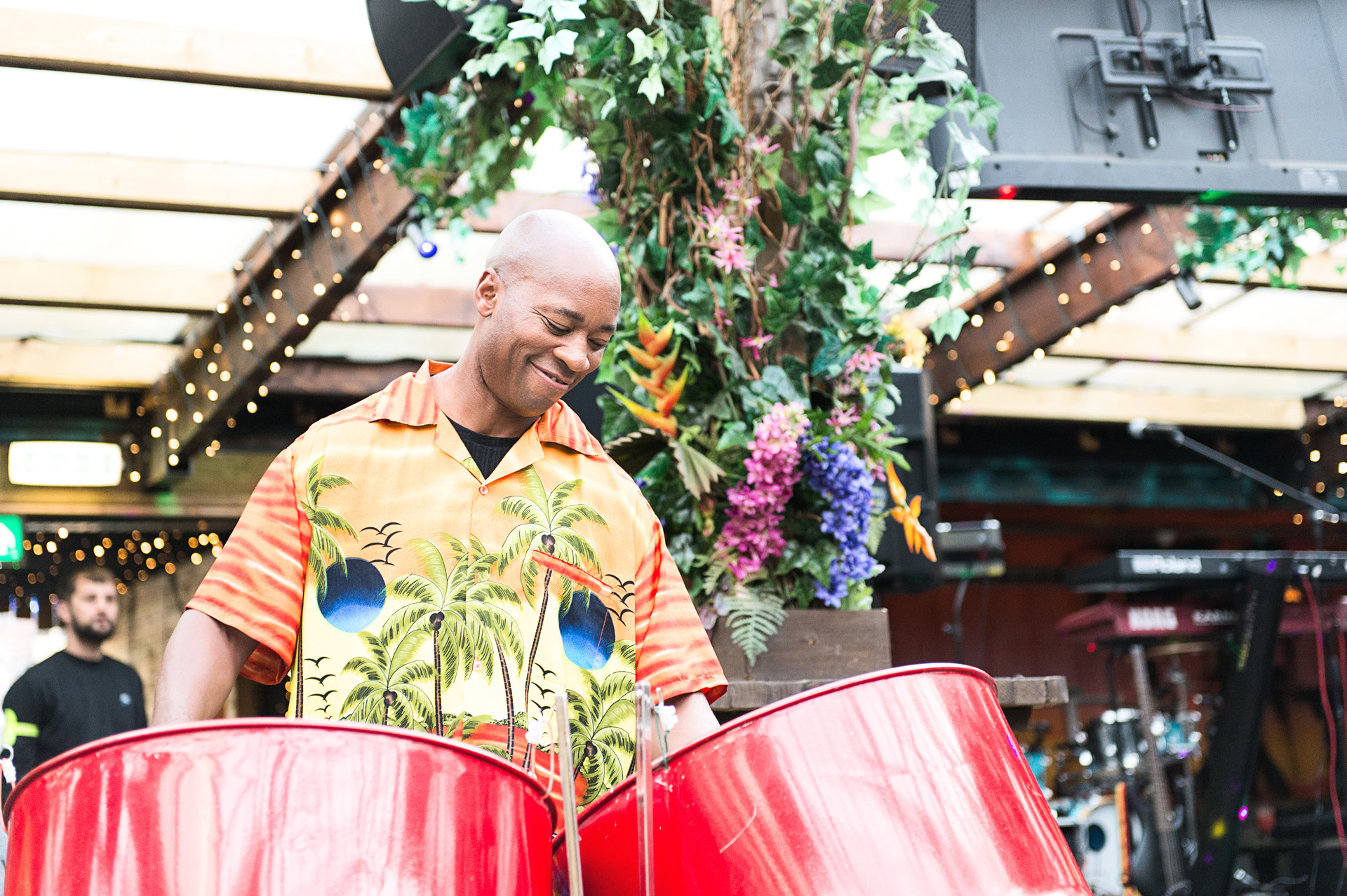 A steel drummer performing beneath hanging greenery and festoon lights at a summer party
