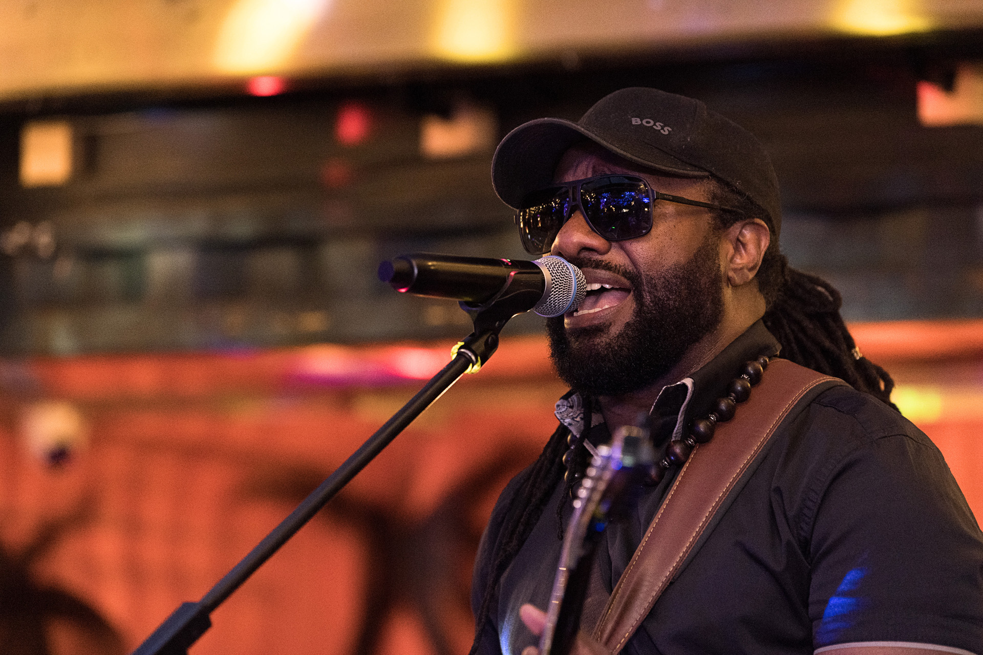 A singer in a cap and sunglasses performing into a microphone with a guitar