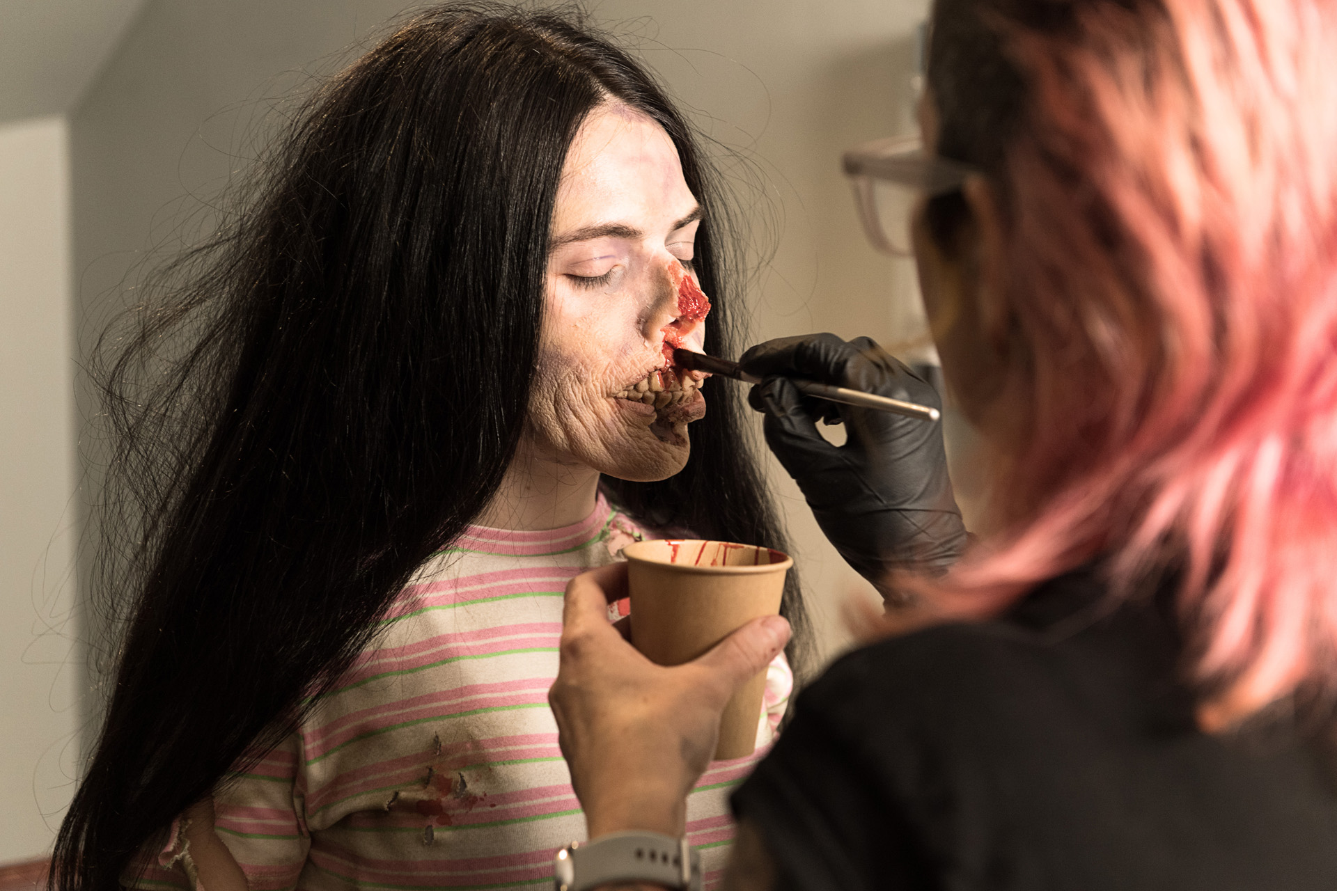 A special effects artist applying zombie makeup to a performer holding a cup
