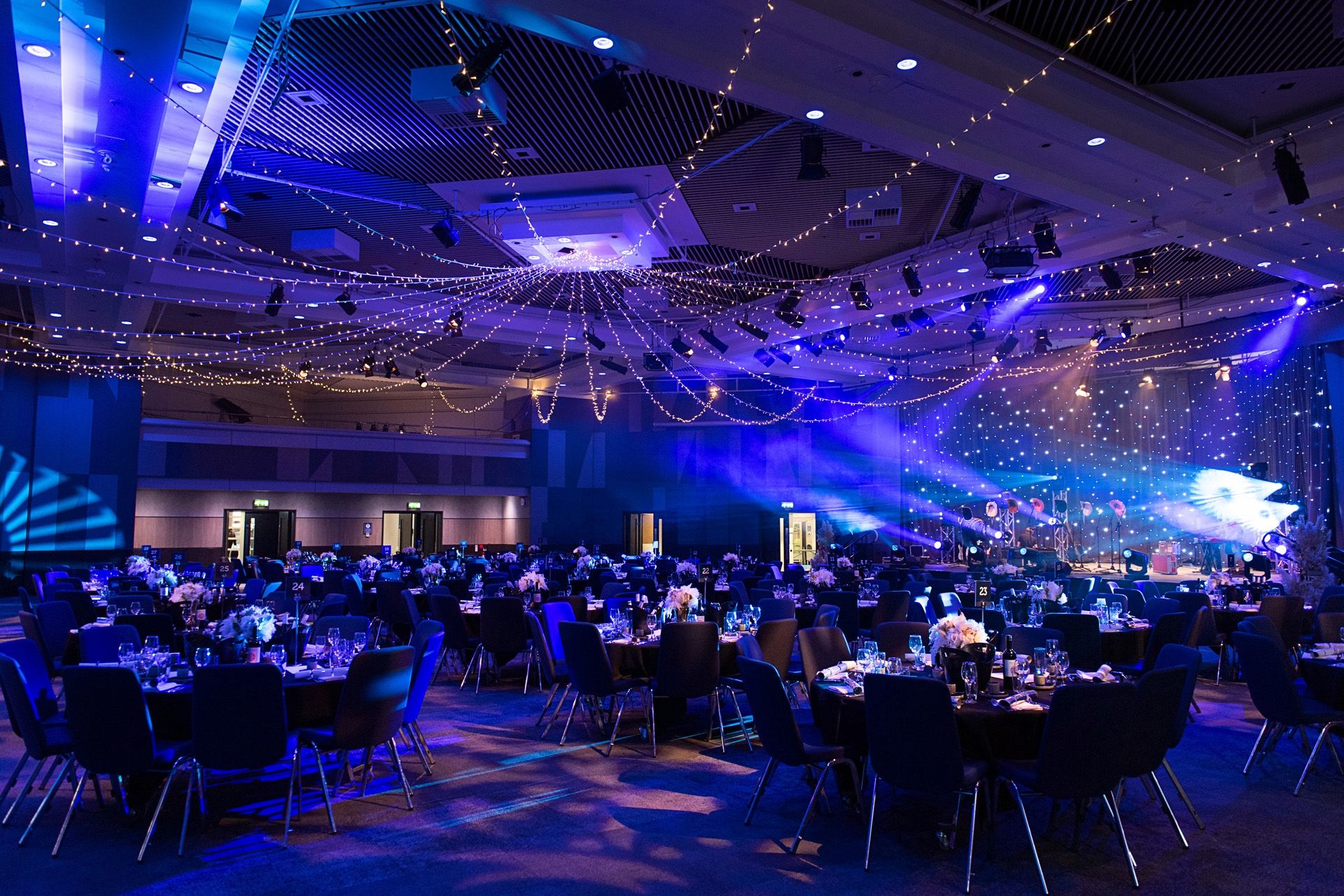 A ballroom laid with round dinner tables under blue lighting and draped fairy lights