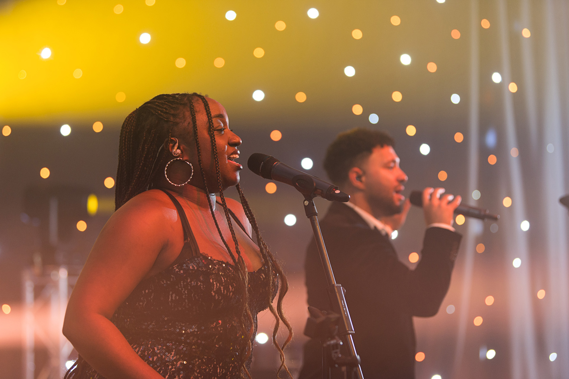 Two singers performing side by side in front of a curtain of warm lights
