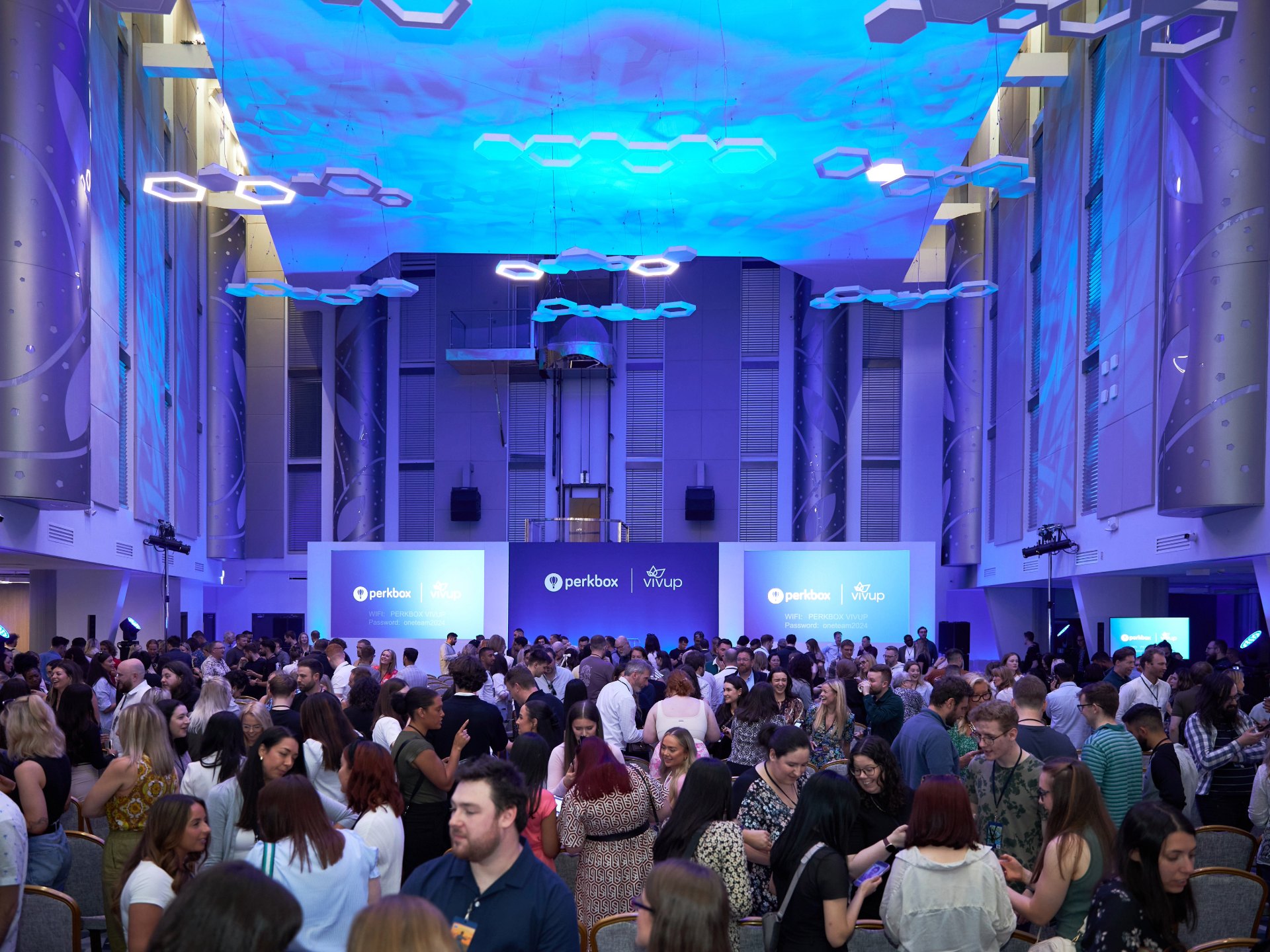 A large standing audience filling a hall beneath a blue ceiling installation and stage