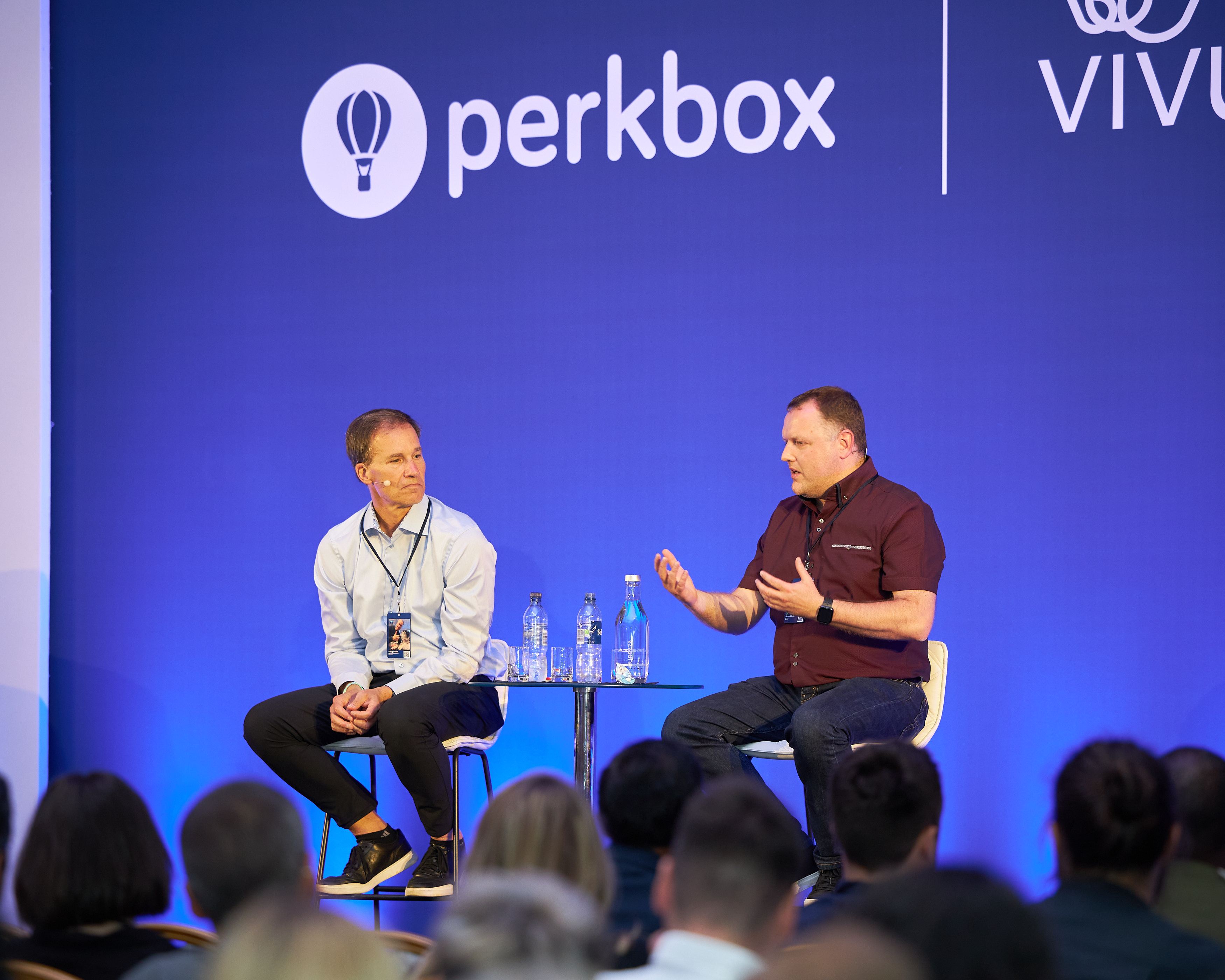An audience watching two speakers seated on stage below the merged company logos