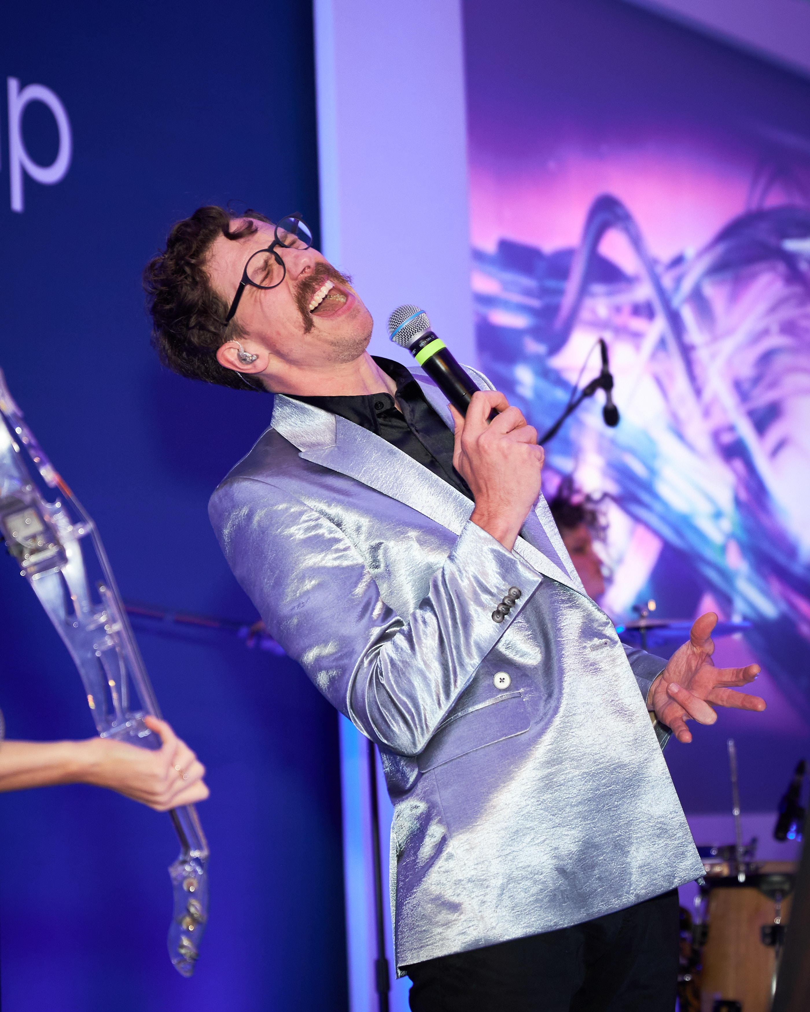 A performer in a silver jacket singing while a violinist plays behind him