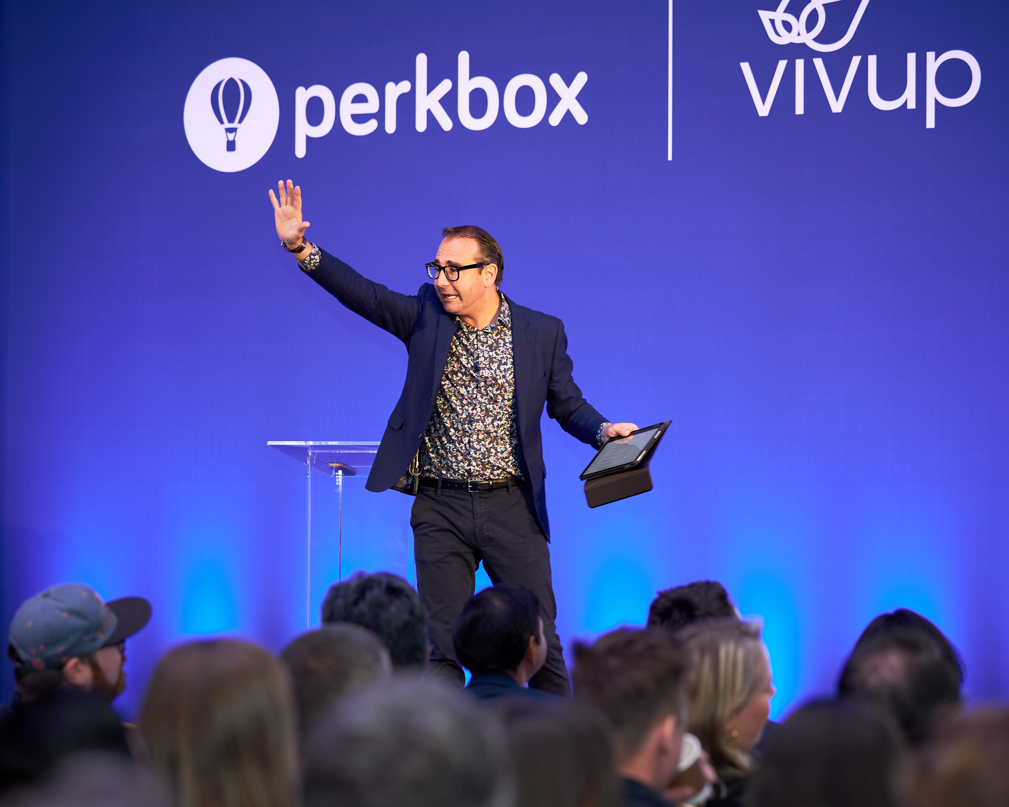 A speaker waving to the audience on stage below two company logos