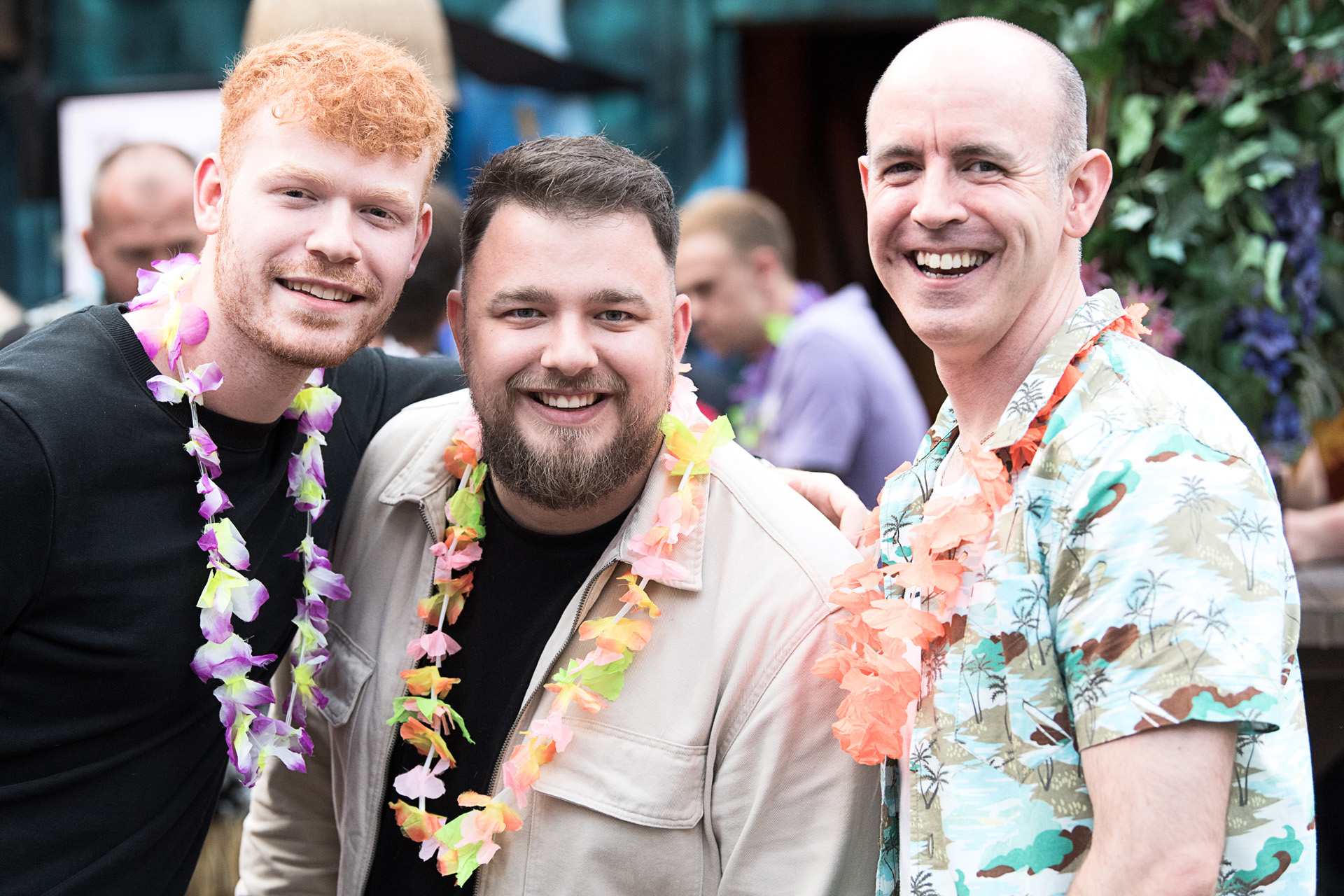 Three colleagues in flower garlands smiling together at a tropical themed party