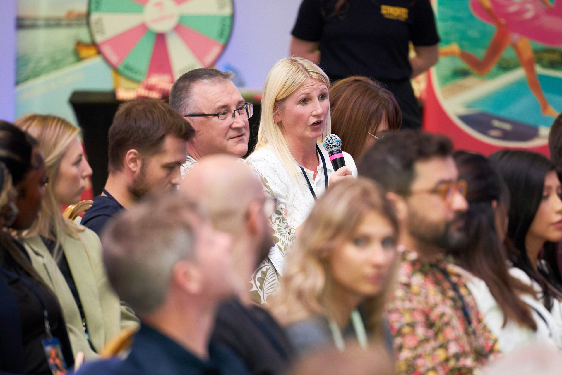 A woman with a microphone addressing colleagues beside a colourful prize wheel