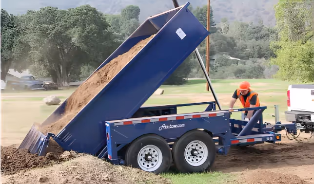 An Air-Tow hydraulic dump trailer in action, tilted at a high angle to unload a full load of soil at a worksite.
