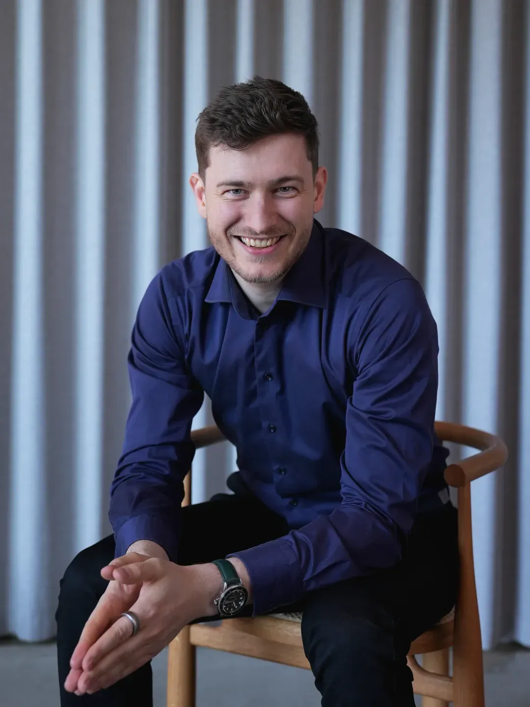 Smiling young man with short brown hair, wearing a dark blue shirt and black pants, sitting on a wooden chair with grey curtains in the background.