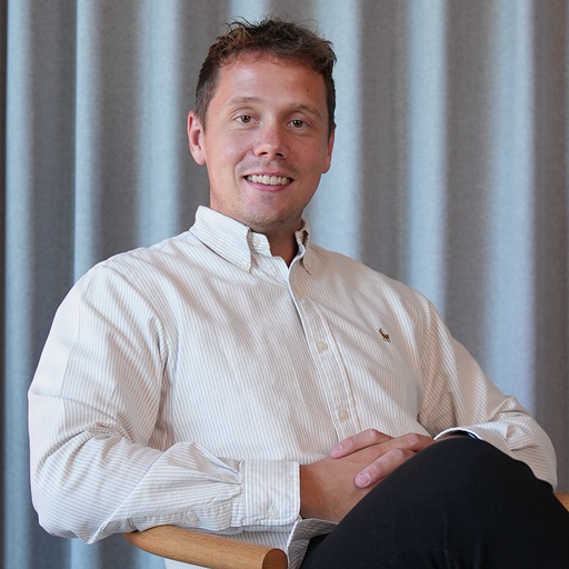 Smiling man with short curly hair wearing a light striped button-up shirt sitting with hands clasped on his lap against a gray curtain backdrop.