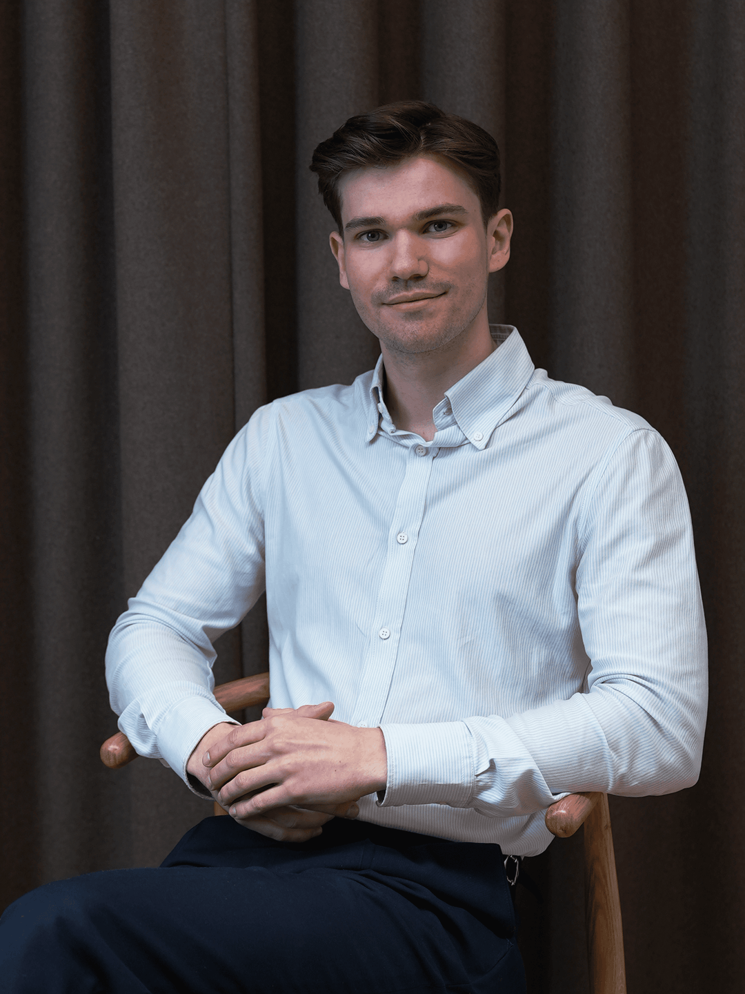 Young man with brown hair wearing a light blue button-up shirt sitting on a wooden chair against a dark curtain background.