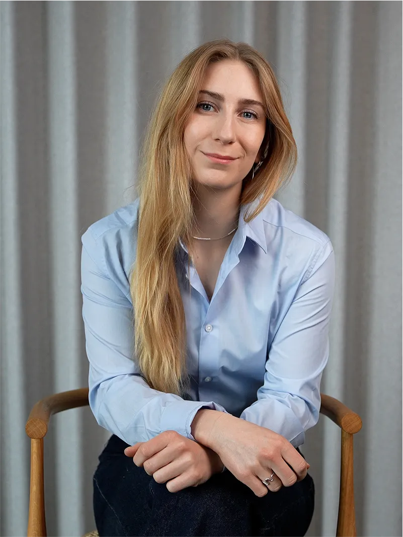 Young woman with long blonde hair wearing a light blue button-up shirt, sitting on a wooden chair with a neutral gray curtain background.