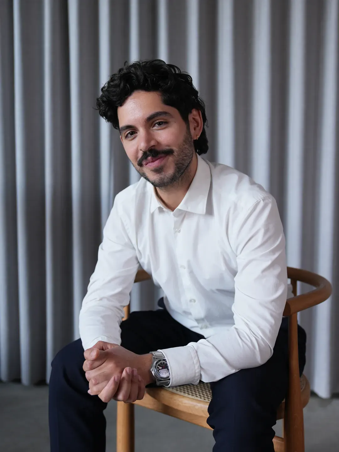 Man with curly hair and beard sitting on a wooden chair wearing a white shirt and dark pants, smiling at the camera.