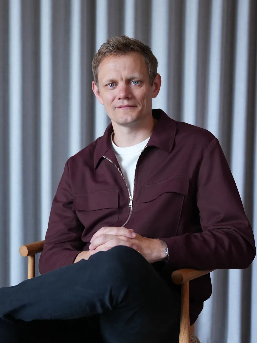 Man with short light brown hair wearing a maroon zip-up jacket and white shirt sitting on a wooden chair with legs crossed in front of a gray curtain.
