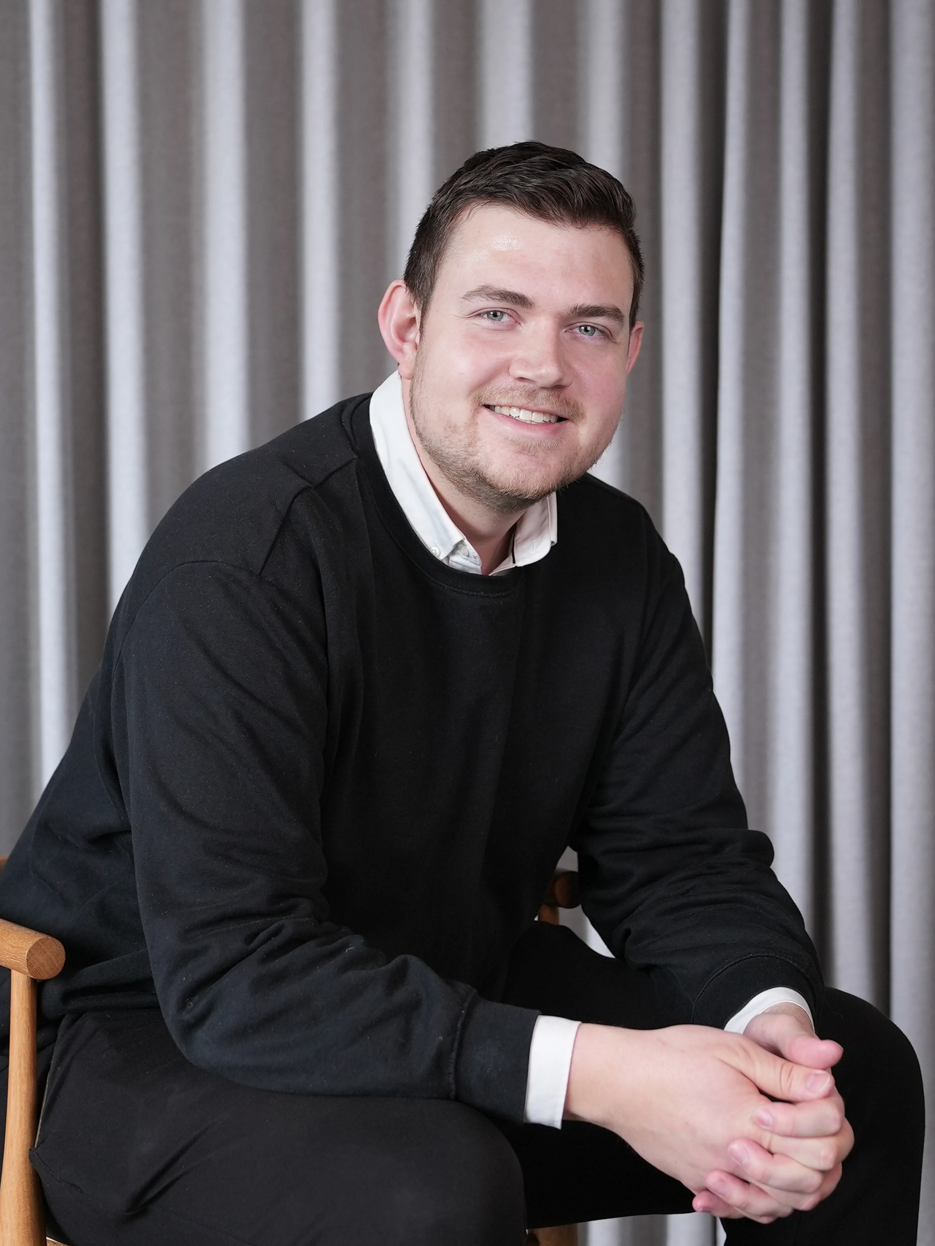 Smiling man with short dark hair wearing a black sweater over a white collared shirt, sitting on a wooden chair with clasped hands in front of gray curtains.