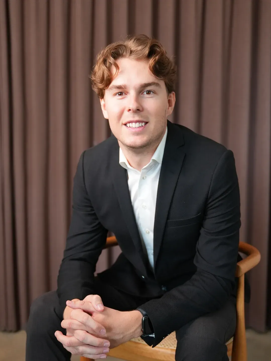 Young man in a black suit and white shirt sitting on a wooden chair with hands clasped, smiling at the camera.