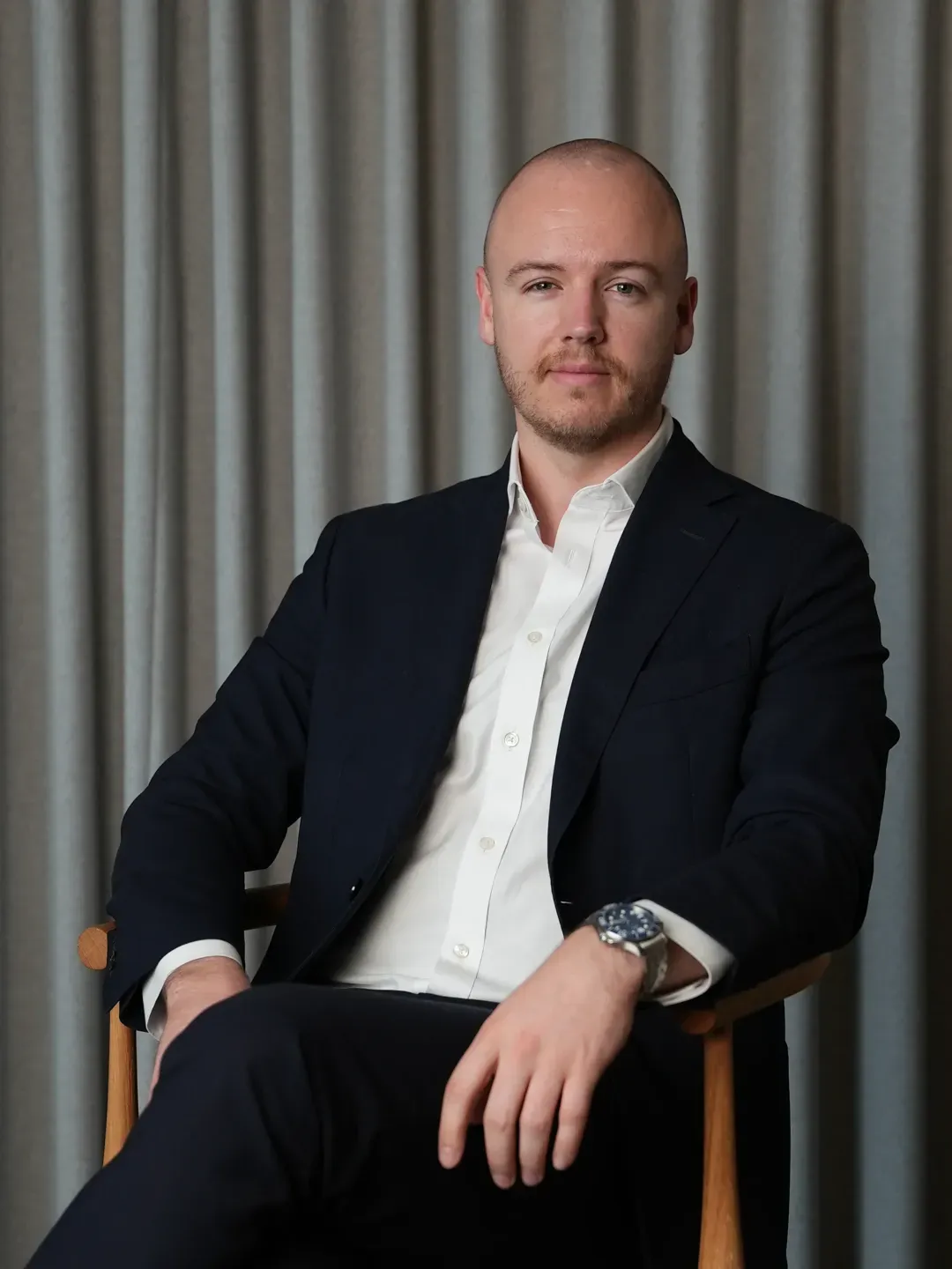 Man with a shaved head and beard wearing a black suit and white shirt sitting on a wooden chair against a gray curtain background.
