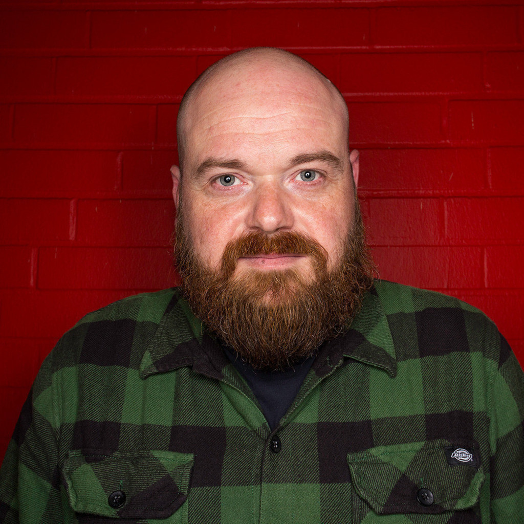 Bald man with a full beard wearing a green and black checkered shirt standing against a red brick wall.