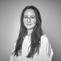 Portrait of a smiling woman with long hair and glasses wearing a light-colored blouse against a plain background.