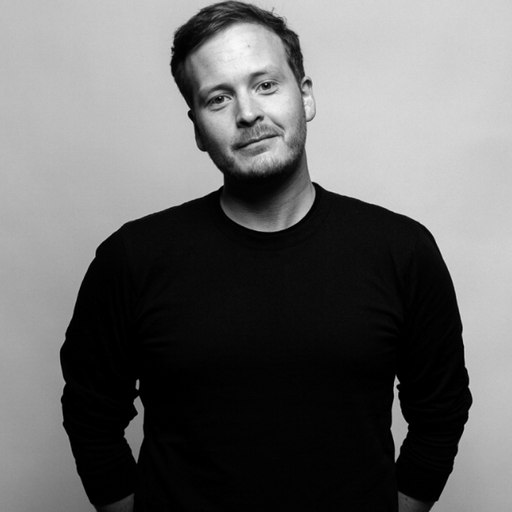Black and white portrait of a man with short hair and a beard, wearing a black long-sleeve shirt, standing against a plain background.