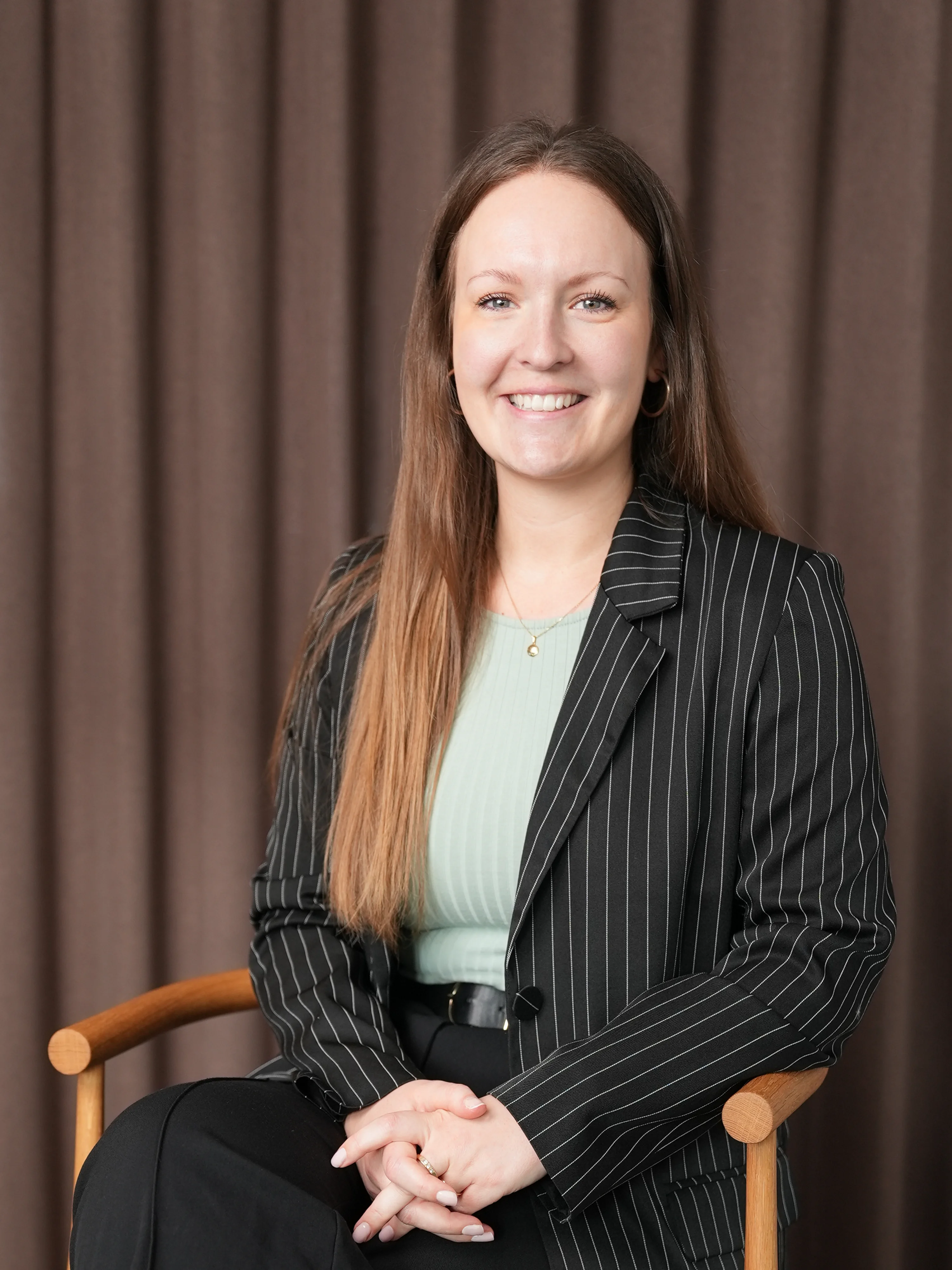 Smiling woman with long brown hair wearing a black pinstripe blazer and mint green top, sitting on a wooden chair against a brown curtain background.