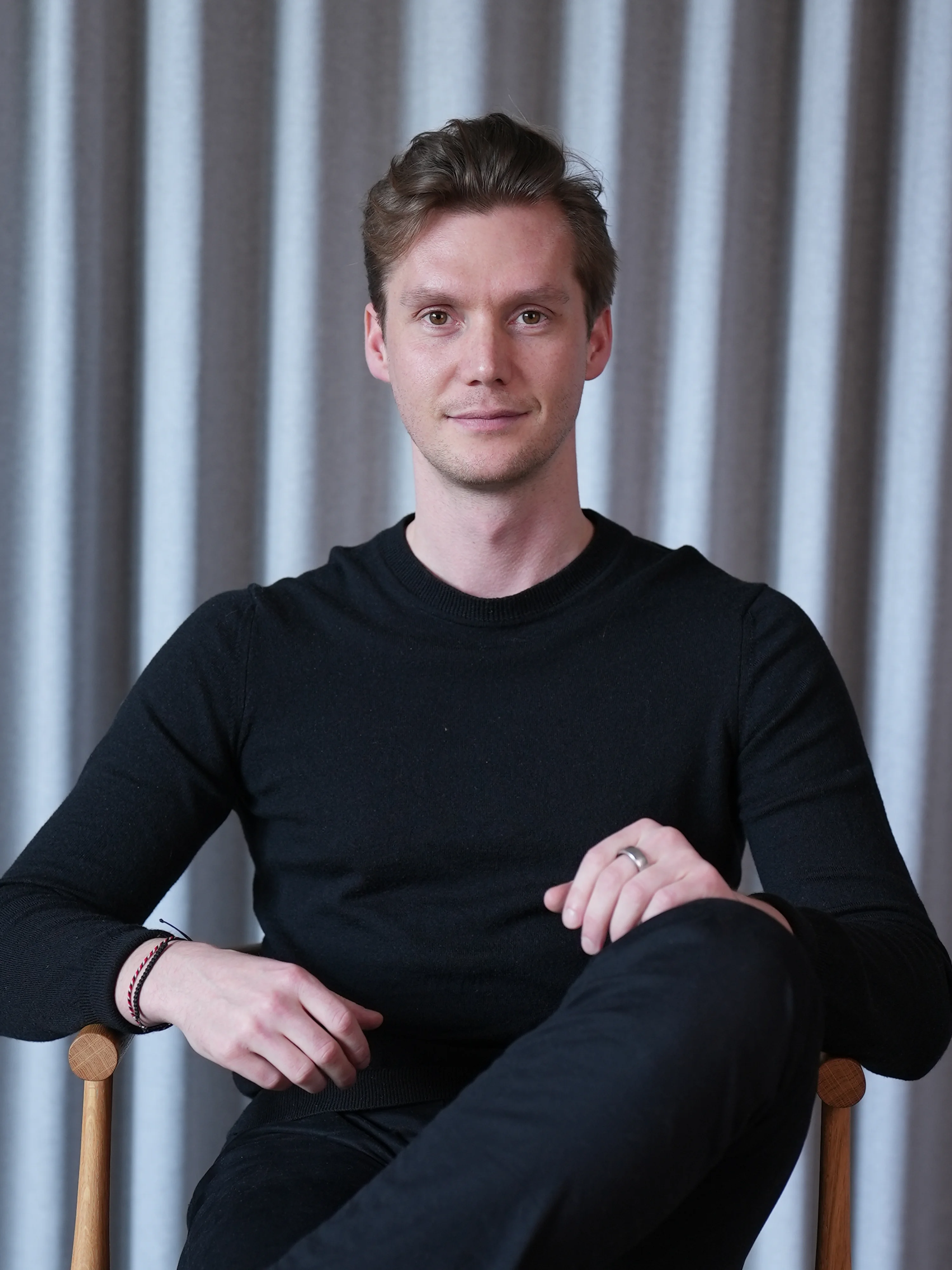 Young man with short brown hair sitting on a wooden chair, wearing a black sweater and black pants, in front of a grey curtain.