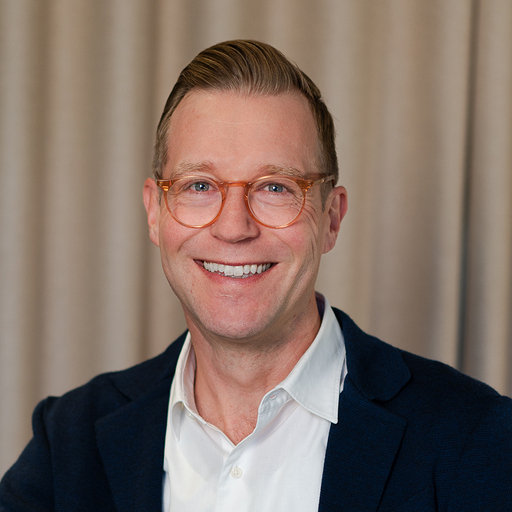 Smiling man with short light brown hair and orange glasses wearing a white shirt and dark blazer in front of beige curtains.