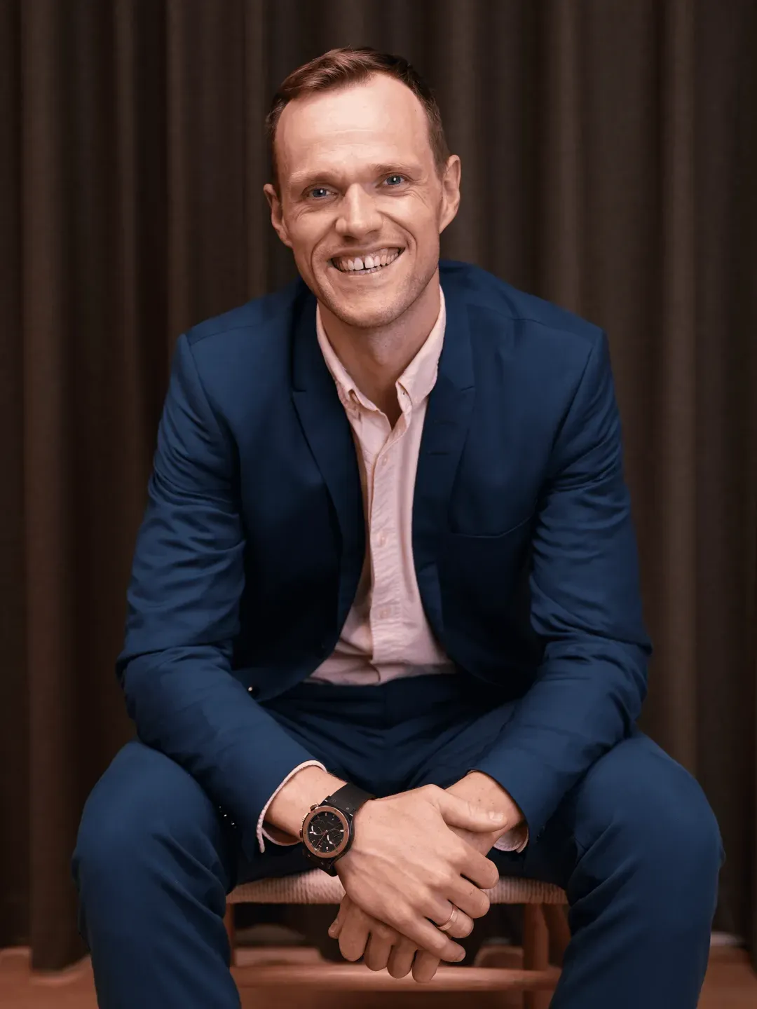 Smiling man in a blue suit and light pink shirt sitting with hands clasped wearing a black wristwatch.