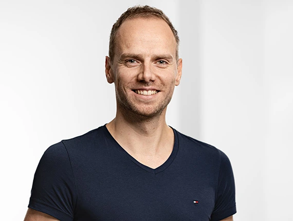 Smiling man with short hair wearing a dark blue V-neck t-shirt against a plain light background.