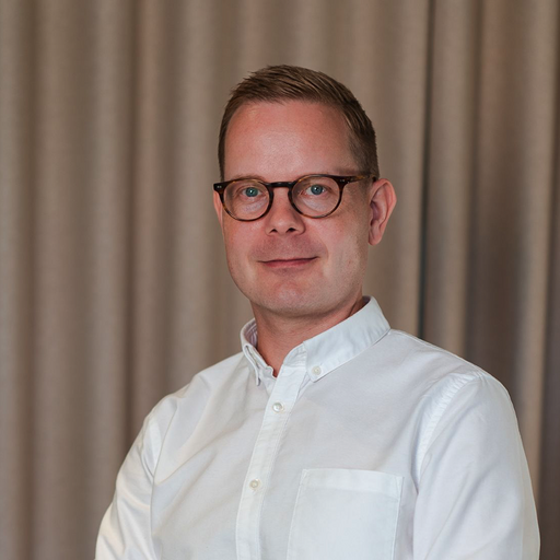 Man with short light brown hair and glasses wearing a white button-up shirt, standing in front of beige curtains.