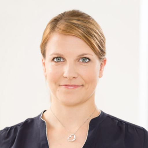 Portrait of a woman with light brown hair pulled back, wearing a dark top and a heart-shaped pendant necklace, smiling gently.
