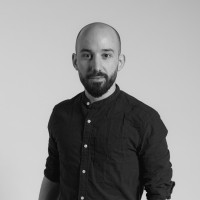 Portrait of a bald man with a beard wearing a dark button-up shirt against a plain background.