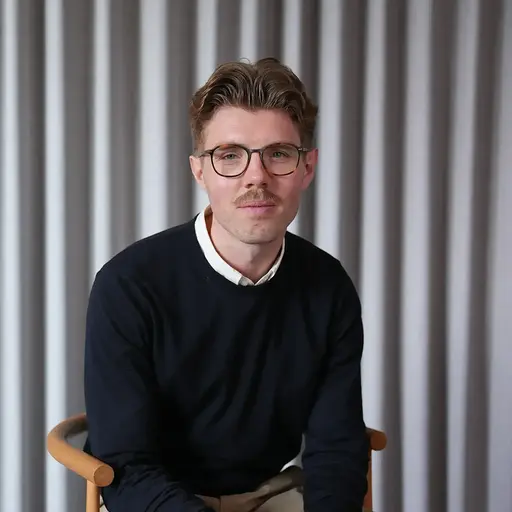 Man with glasses and mustache wearing a black sweater sitting on a wooden chair in front of a gray curtain.