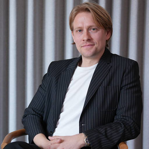 Man with light brown hair wearing a black pinstripe blazer and white t-shirt sitting on a wooden chair against a grey curtain background.