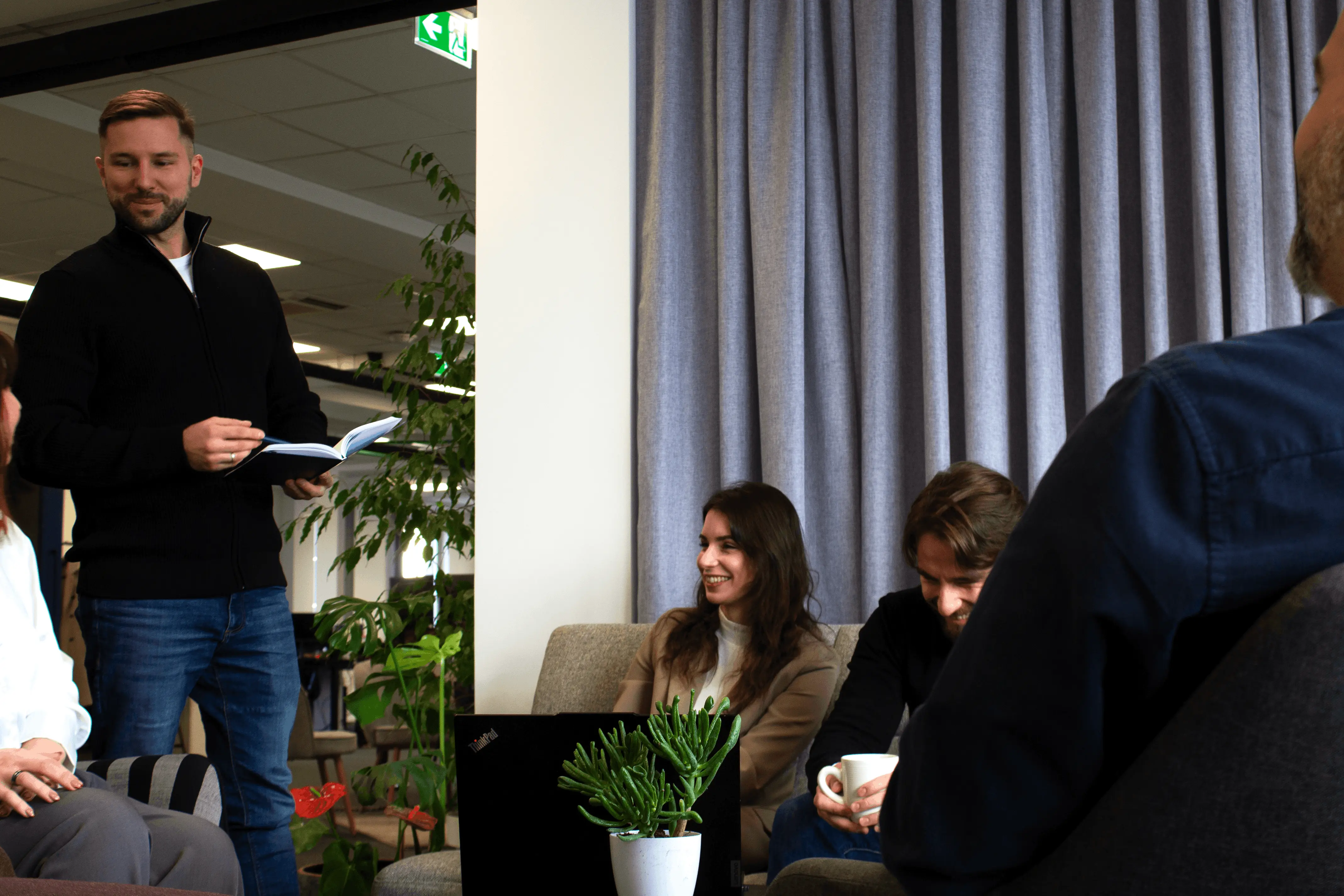 Group of four people in a casual office setting, one standing with a notebook while others sit and smile.